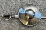 Hammered Steel Buckler Shield, 14 inch - Man At Arms displayed with a sword on a stone surface.