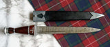 Scottish Long Dirk with Damascus blade and ebony grip alongside its sheath on tartan fabric.