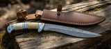Replica Persian Fighting Knife with Damascus Blade Man At Arms on a wooden surface with a leather sheath.