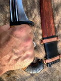 Close-up of hand gripping the hilt of an Iberian falcata sword with a decorative scabbard in the background.