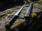 Late Crusader medieval sword with hand forged blade displayed beside its sheath on mossy stone.