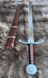 The Bedford Medieval War Sword displayed with a leather scabbard on a textured surface.