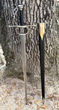 Sidesword With Complex Hilt by Kingdom Of Arms next to its leather scabbard, set against a tree backdrop.