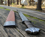 The Saxon Langsax or Long Seax displayed with its scabbard on a wooden surface in a forest setting.